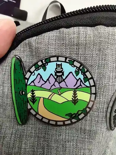 A close-up photo shows a circular enamel pin attached to a gray fabric bag, possibly a backpack. The pin depicts a stylized…