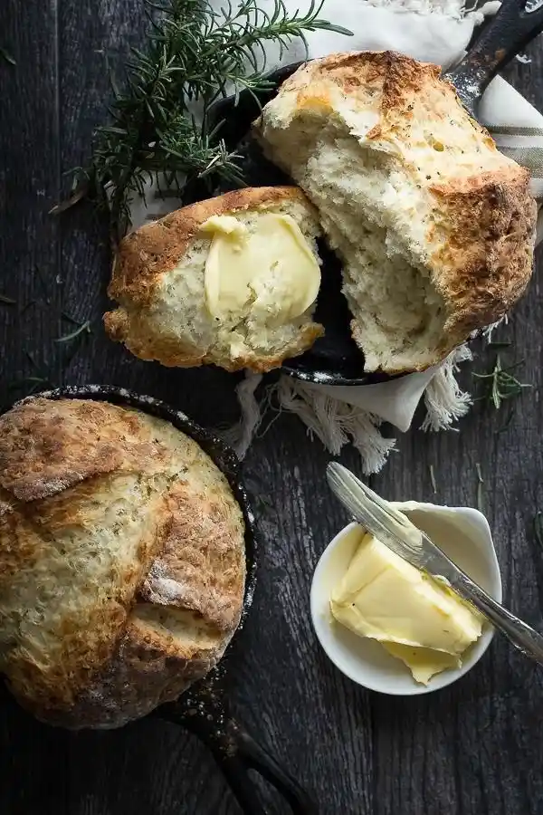 A rustic, golden-brown loaf of bread sits in a cast-iron skillet, with another loaf sliced open to reveal soft, airy crumb…