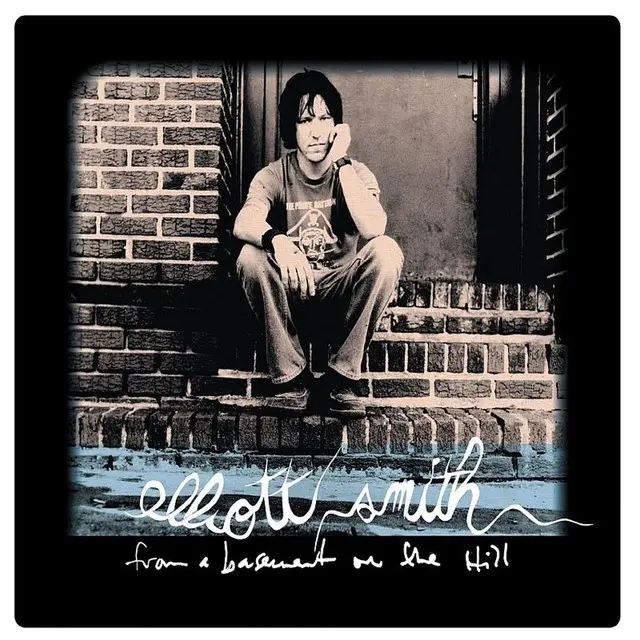 A black-and-white photo of Elliott Smith sitting on brick steps, looking pensive, with handwritten-style text at the bottom…