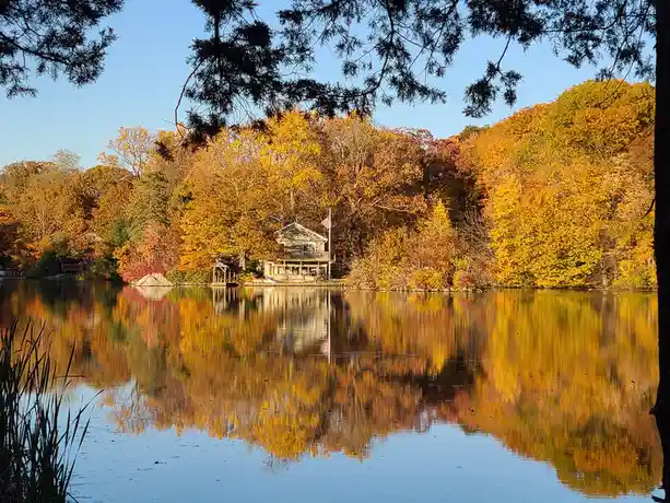 A serene autumn landscape shows a lake reflecting vibrant yellow and orange trees, with a wooden cabin on the shore under a…