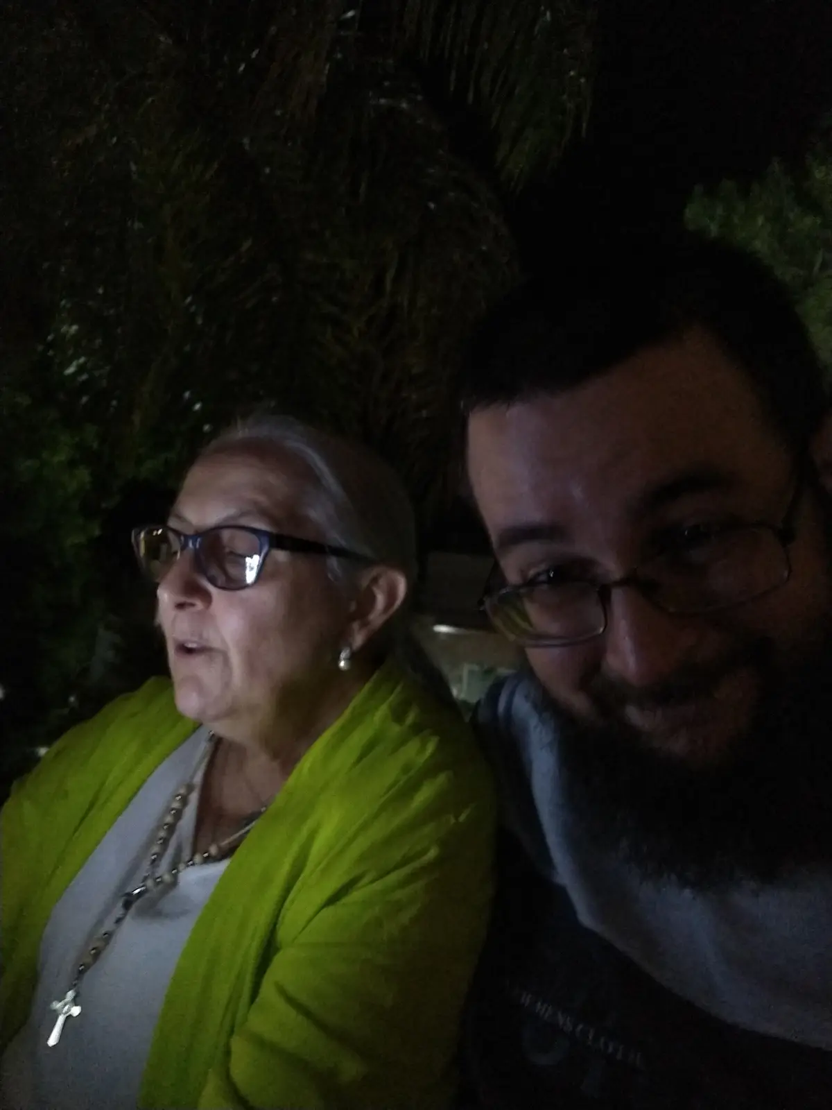 A low-light selfie shows a man with glasses and a beard smiling at the camera while sitting next to an older woman with…