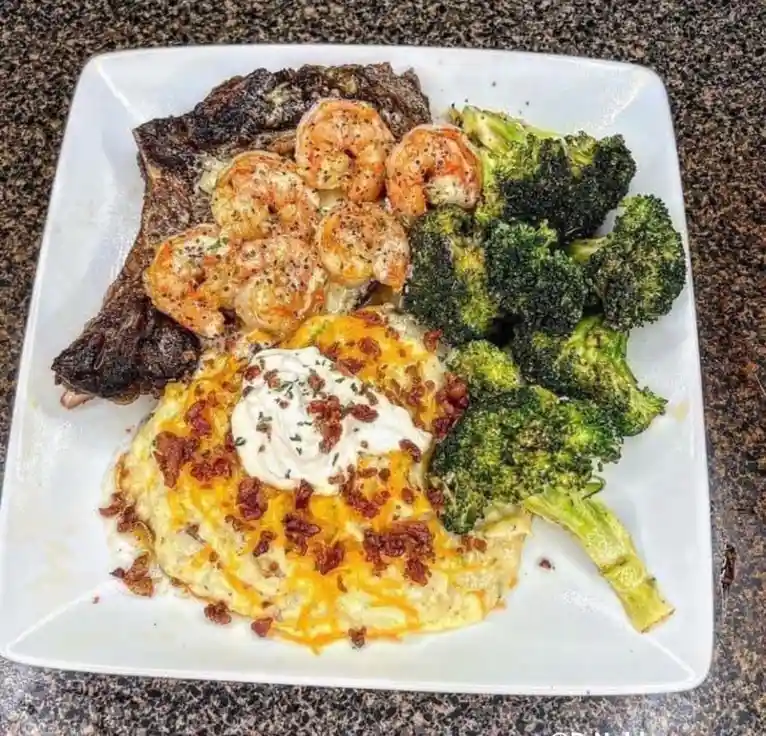 A white plate holds a large, heavily charred steak, a pile of cooked shrimp, a serving of roasted broccoli, and a cheesy…