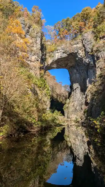 A natural rock arch bridge spans a calm river, surrounded by autumn foliage with orange and green leaves. The clear blue sky…