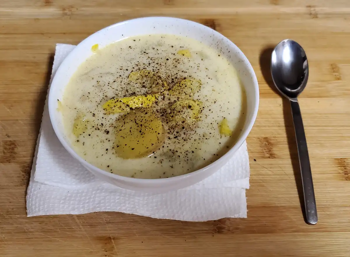 A white bowl filled with creamy soup, topped with black pepper and yellow butter, sits on a white paper towel on a wooden…