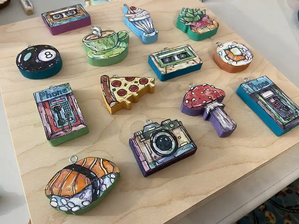 A collection of hand-painted wooden charms shaped like various objects including cassette tapes, a coffee cup, a pizza…