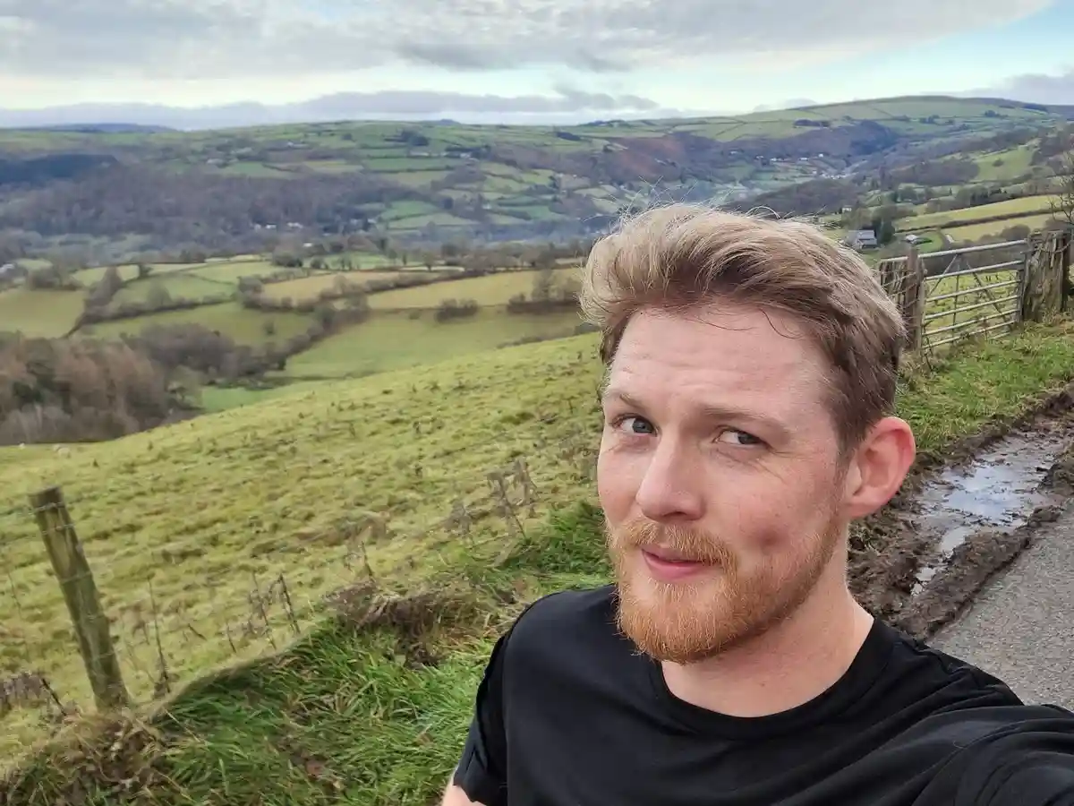 A man with a reddish beard and short hair takes a selfie on a muddy rural path, looking slightly weary or sarcastic at the…