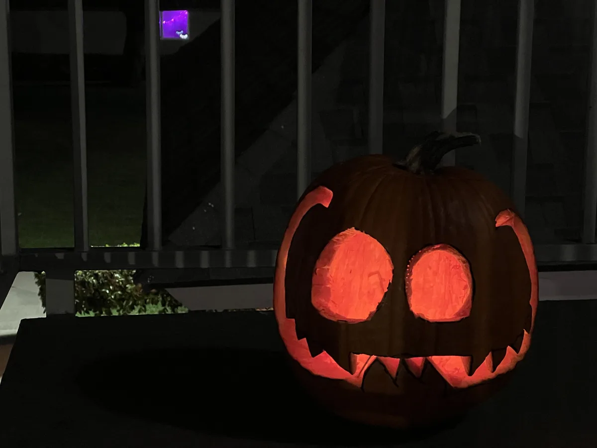 A carved jack-o'-lantern with glowing orange eyes and a jagged grin sits on a dark surface at night, illuminated from…