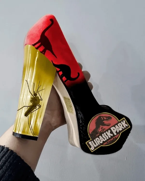 A hand holds a high-heeled shoe with a Jurassic Park logo on the heel and a red upper with black dinosaur silhouettes. The…