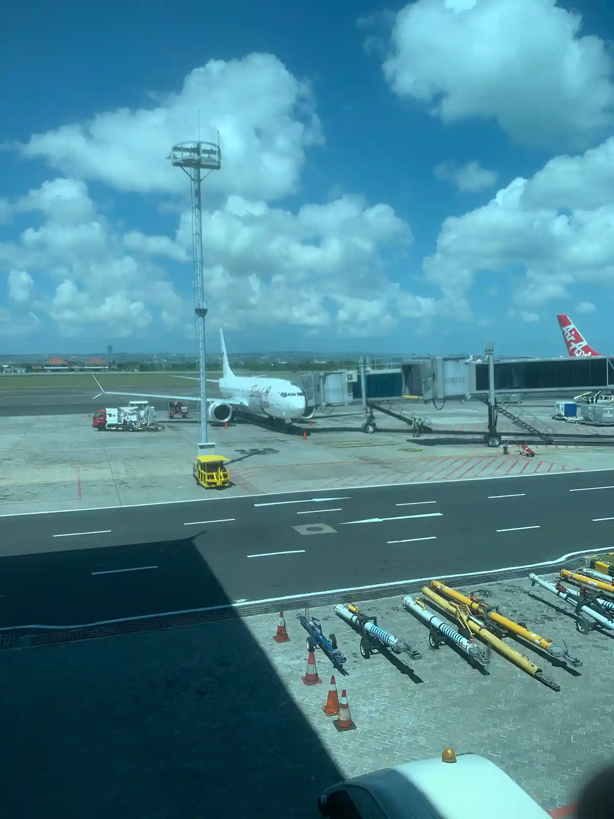 An airport tarmac scene with a white airplane parked at a gate under a partly cloudy blue sky, featuring ground service…