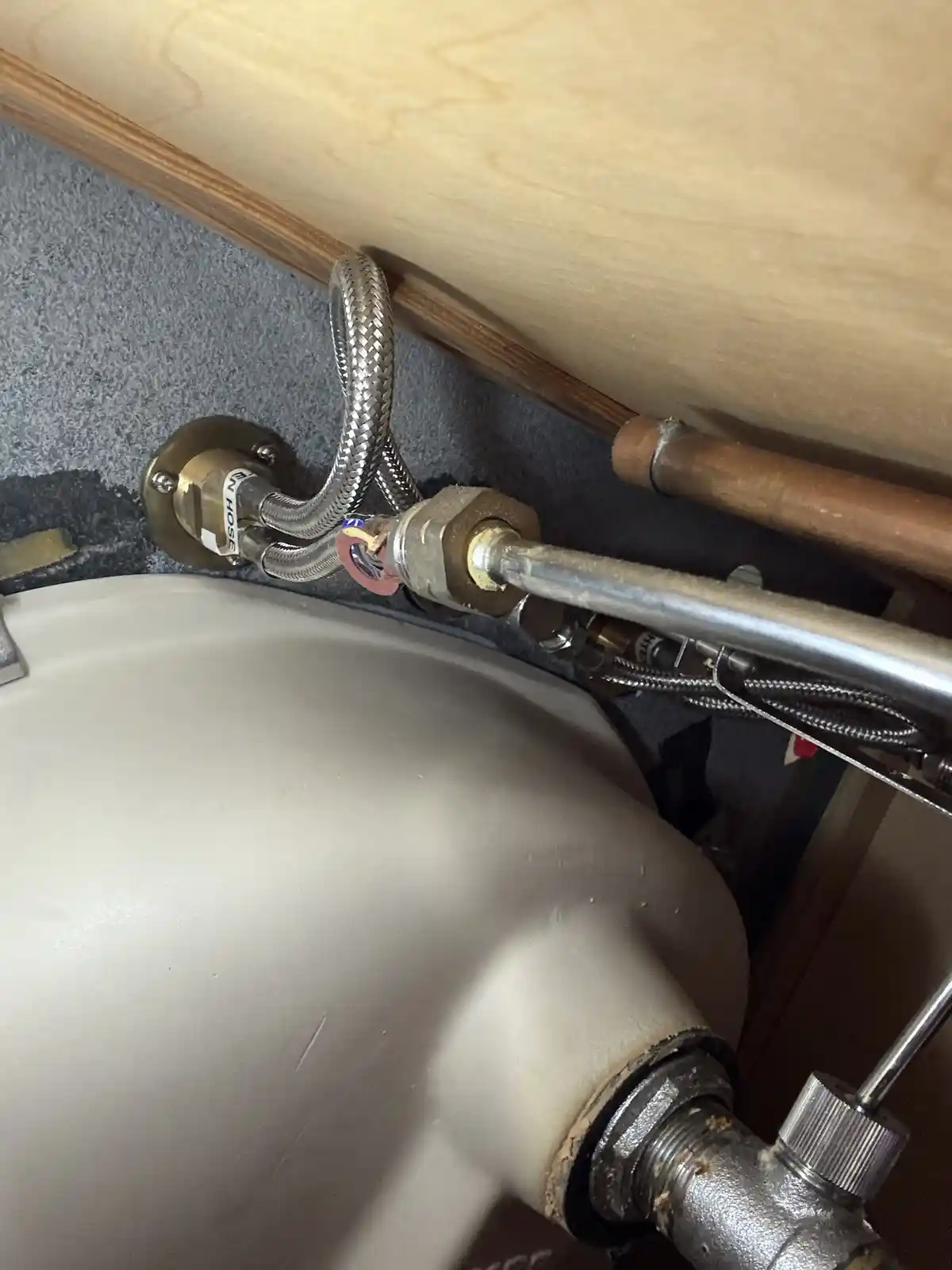 A close-up photo shows plumbing under a sink, with a braided metal hose connected to a brass fitting and a copper pipe…