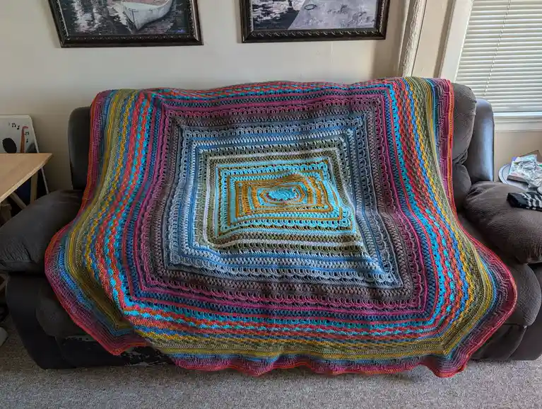 A vibrant, multi-colored crocheted blanket with concentric square patterns is draped over a dark gray couch in a living…