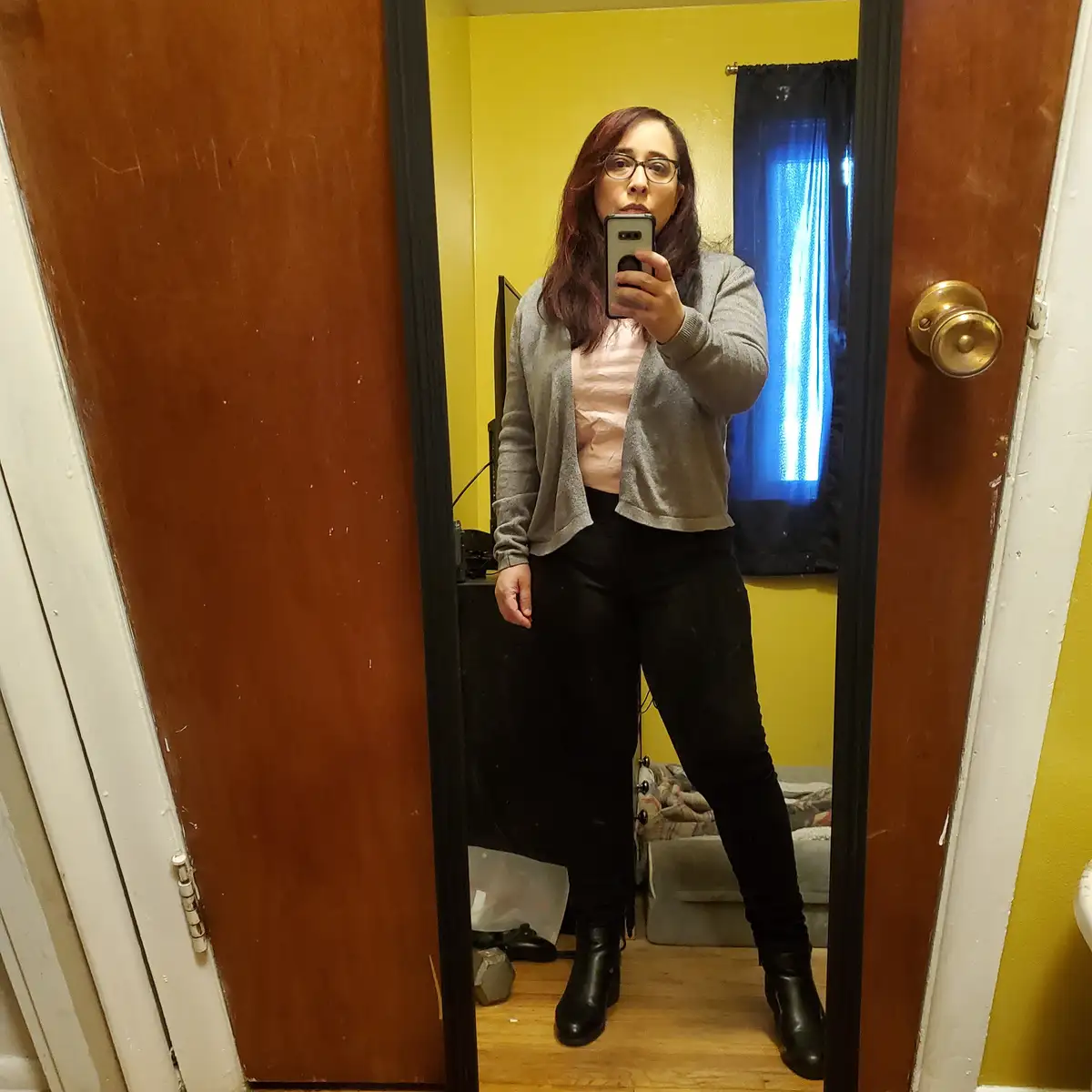 A woman with glasses takes a mirror selfie in a room with yellow walls, wearing a gray cardigan, black pants, and boots…