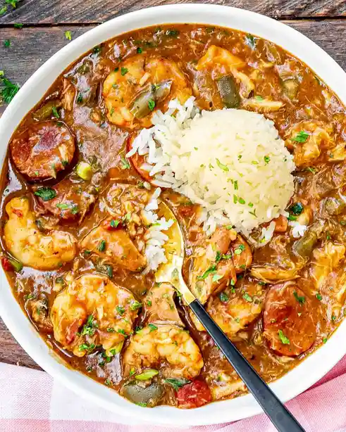 A top-down photo of a white bowl filled with jambalaya, a rich brown stew containing shrimp, chicken, sausage, and…