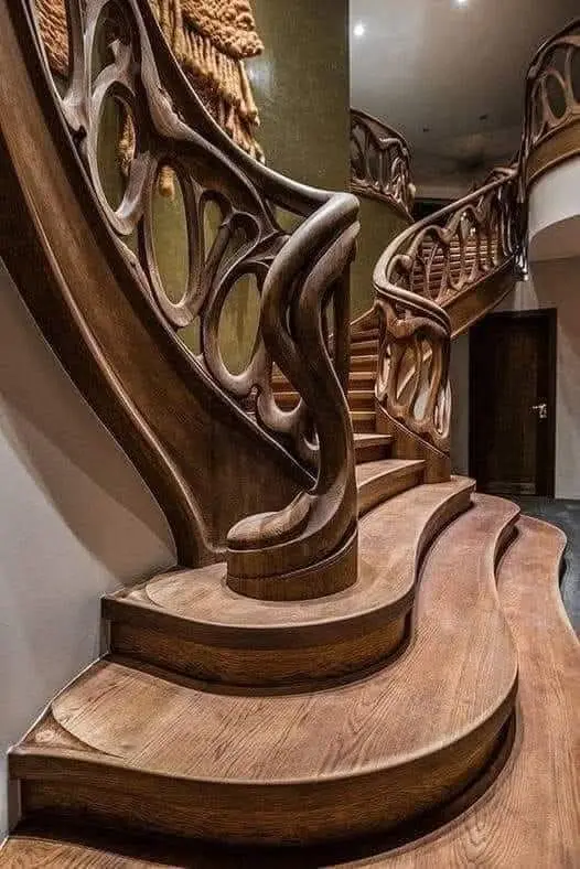 A close-up of an ornate, flowing wooden staircase with intricate carvings on the railing and smooth, curved steps. The image…