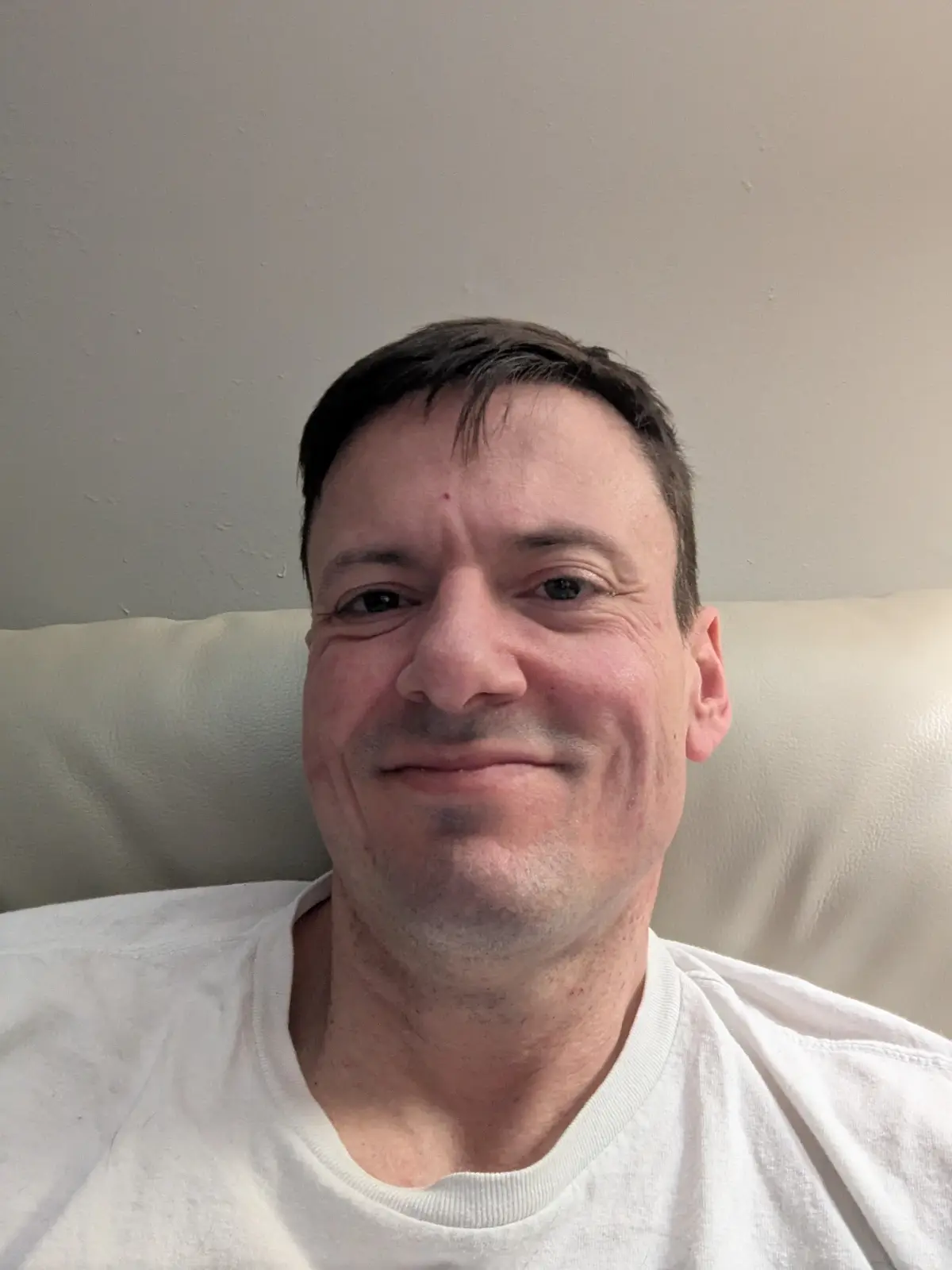 A man with short dark hair and a white t-shirt is sitting on a light-colored couch, looking at the camera with a subtle…