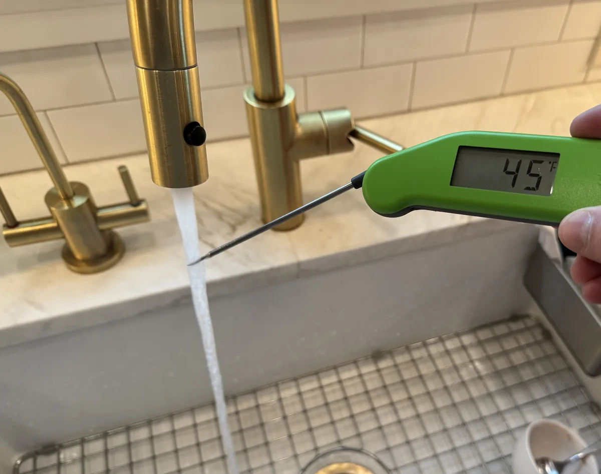 A hand holds a green digital thermometer with its probe inserted into a stream of water flowing from a modern brass faucet…