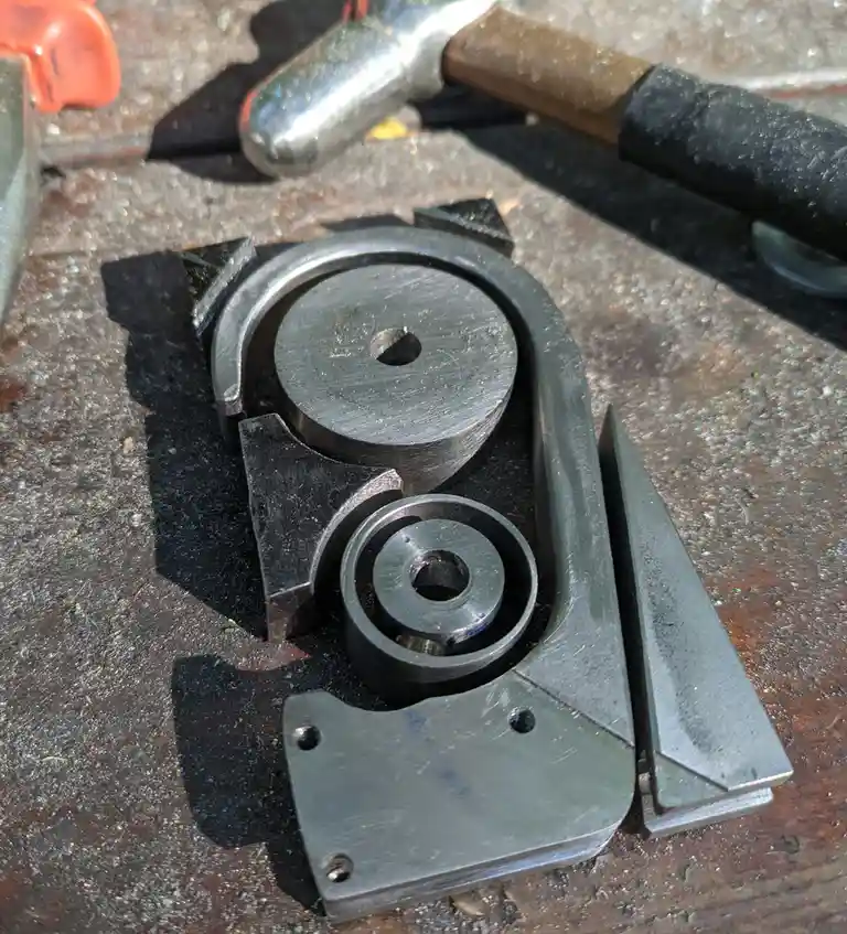 A close-up photo shows a custom-made, complex metal tool with two circular components and angular brackets, resting on a…