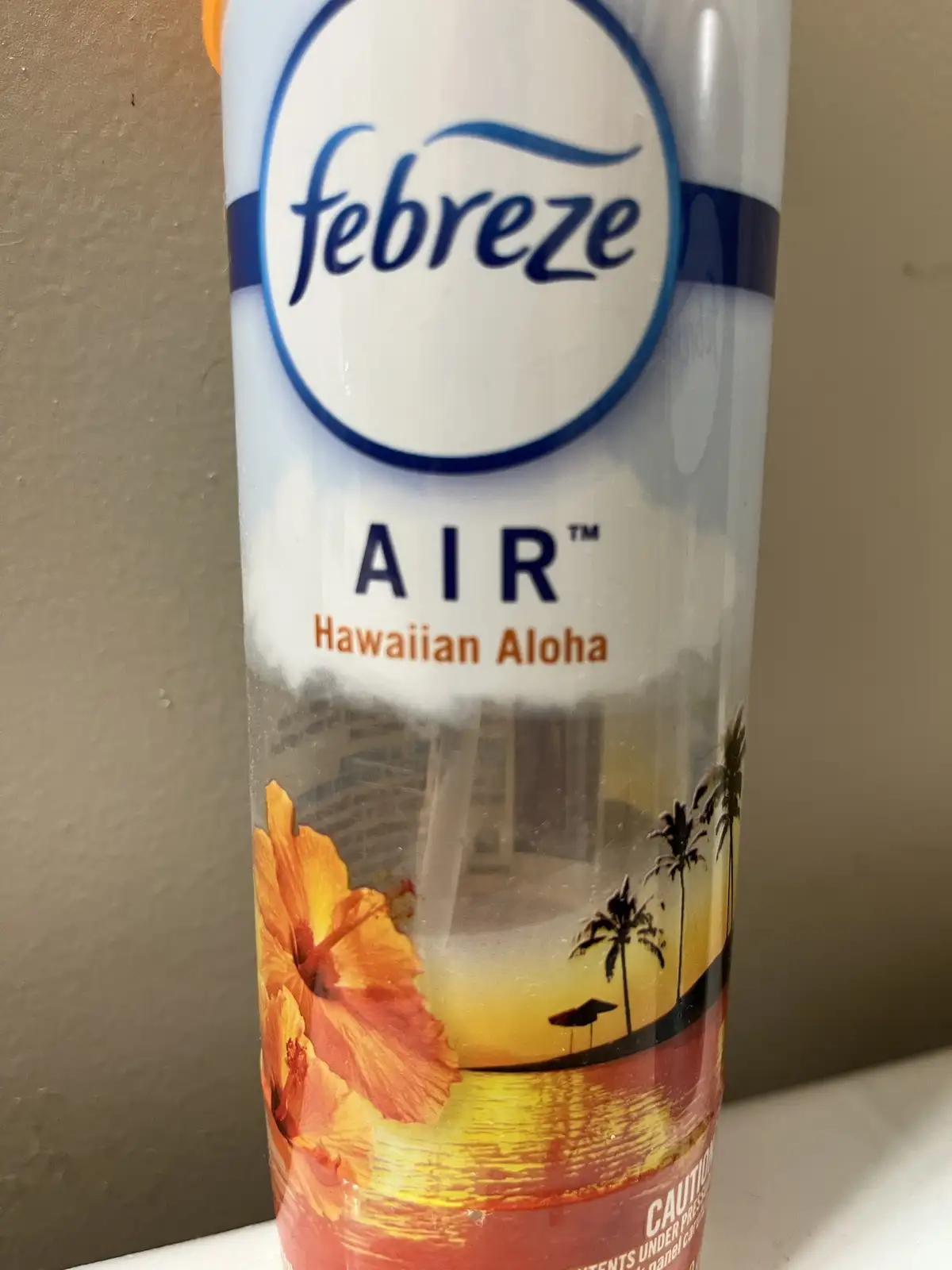 A close-up of a Febreze AIR aerosol can with a Hawaiian Aloha scent, featuring a tropical sunset design with palm trees and…