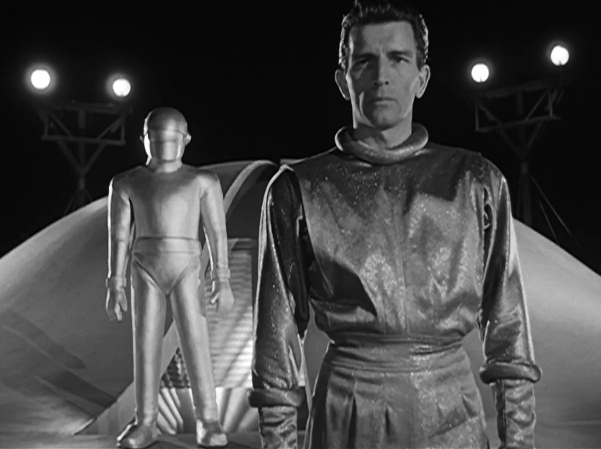 A black-and-white still from a vintage sci-fi film shows a man in a shiny, textured costume standing in the foreground…