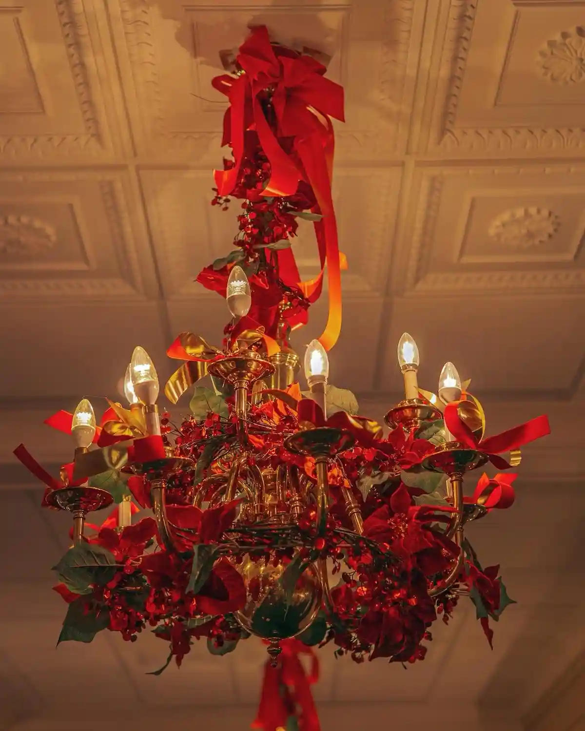 A golden chandelier is heavily decorated with red bows, poinsettia flowers, green leaves, and red berries, transforming it…
