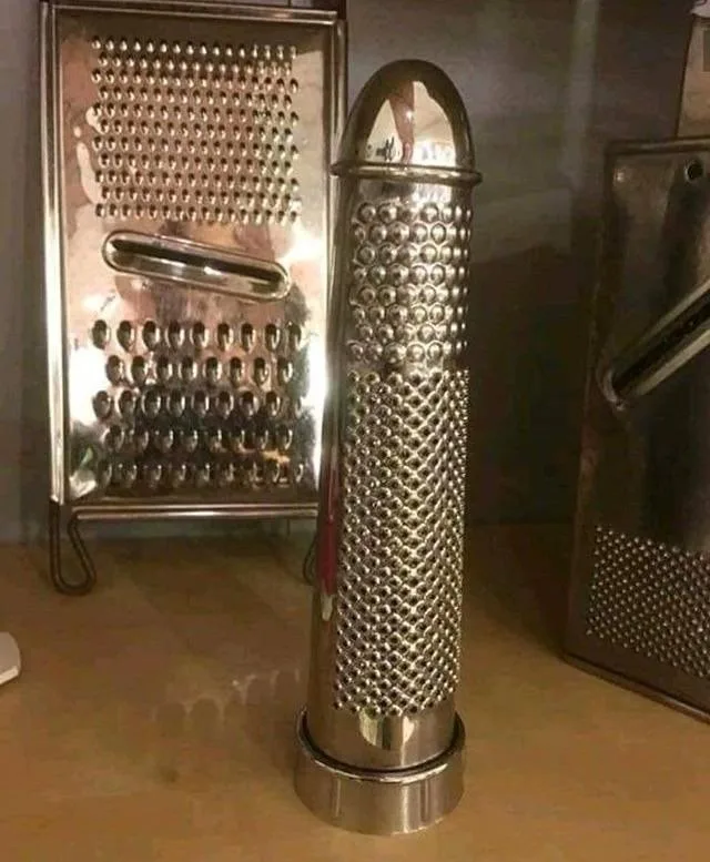 a tall, cylindrical metal grater with a rounded top stands upright on a countertop, flanked by two other flat metal graters…