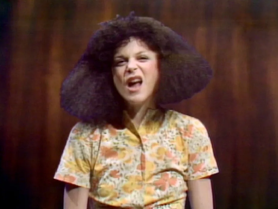 A woman with a large, dark afro hairstyle and a floral-patterned shirt is shown with an exaggerated, angry facial…