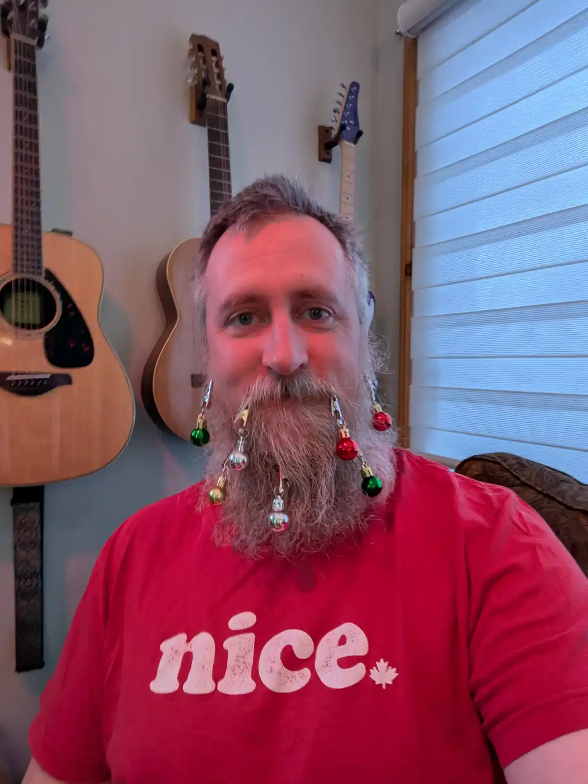 A man with a long grey beard has small red, green, and silver Christmas ornaments clipped to his facial hair, resembling a…