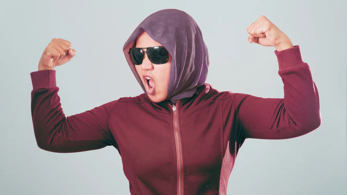 A person wearing a purple hijab and dark sunglasses, flexing both arms with clenched fists while making a loud, open-mouthed…