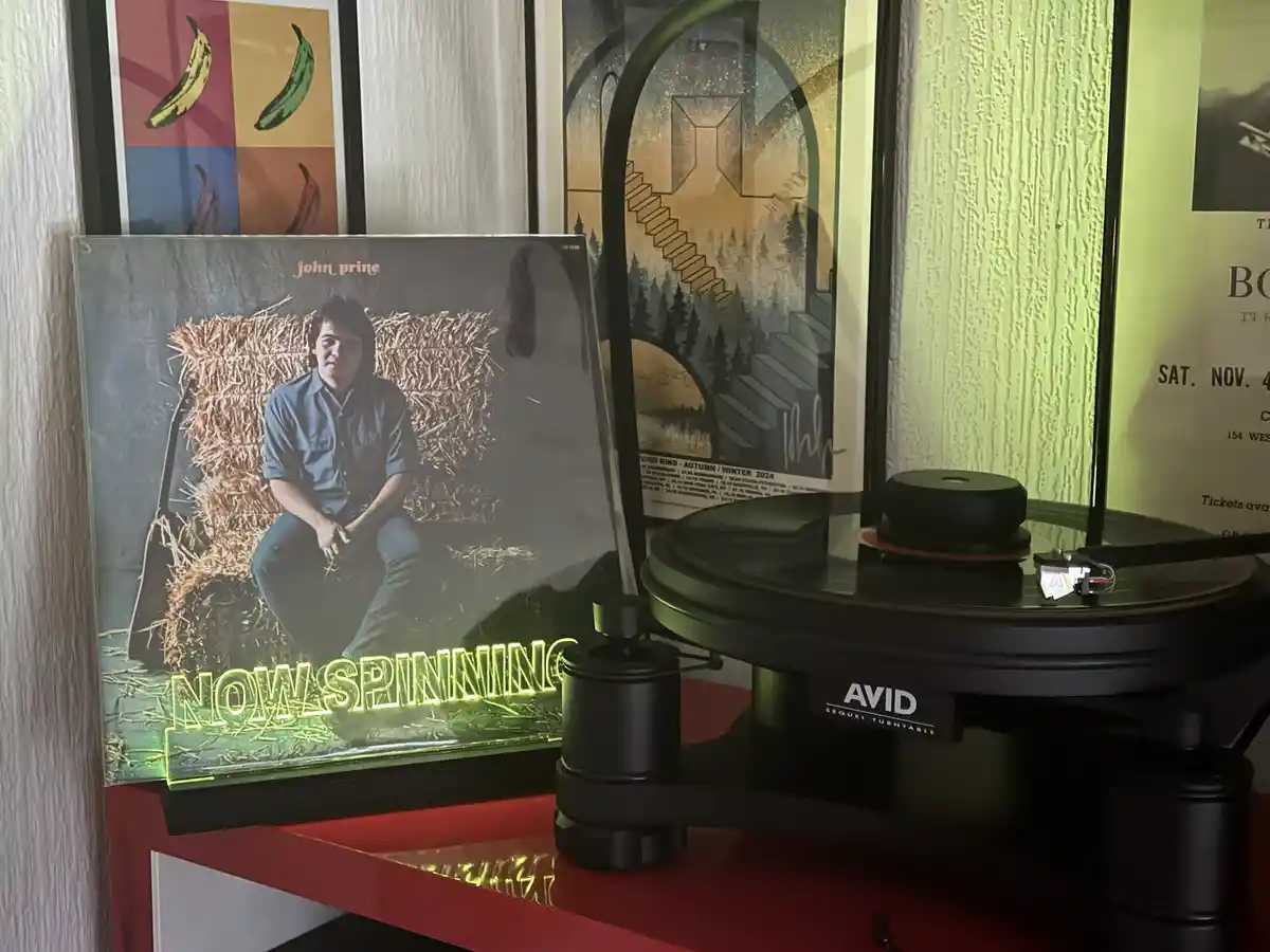 A vinyl record album by John Prine is standing upright next to a black AVID Sequel turntable on a red shelf. The album cover…