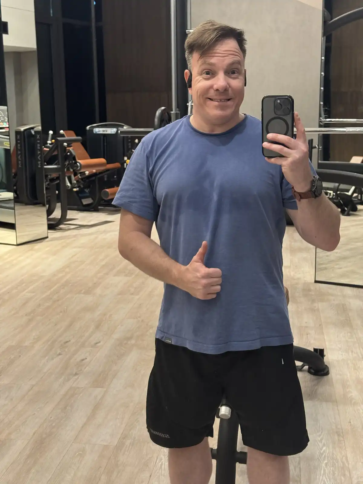 A man in a gym wearing a blue t-shirt and black shorts takes a selfie in a mirror while giving a thumbs-up. He has earbuds…