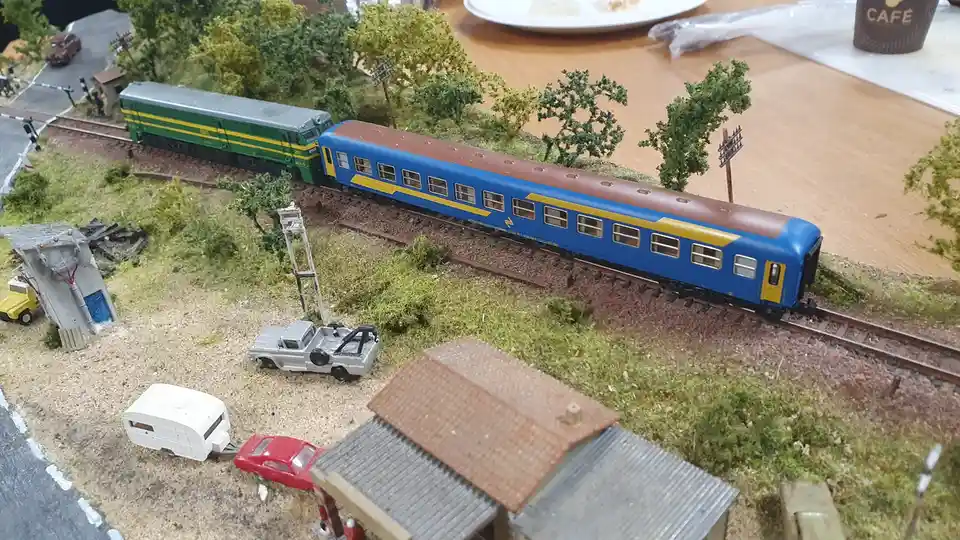 A model train set features a green and yellow locomotive pulling a blue passenger car along tracks through a miniature…