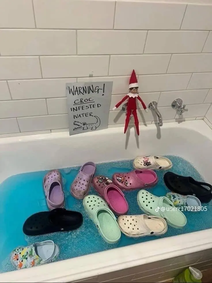 A bathtub filled with soapy blue water and numerous colorful Crocs shoes. A small red elf doll hangs from the faucet, and a…