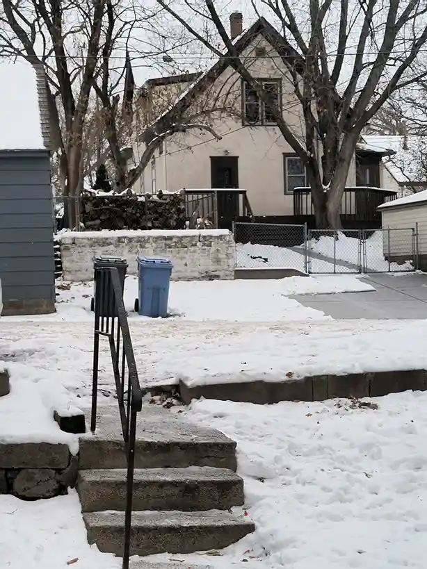 A snowy residential yard with concrete steps leading up to a house, two trash bins, and a fence with stacked firewood. The…