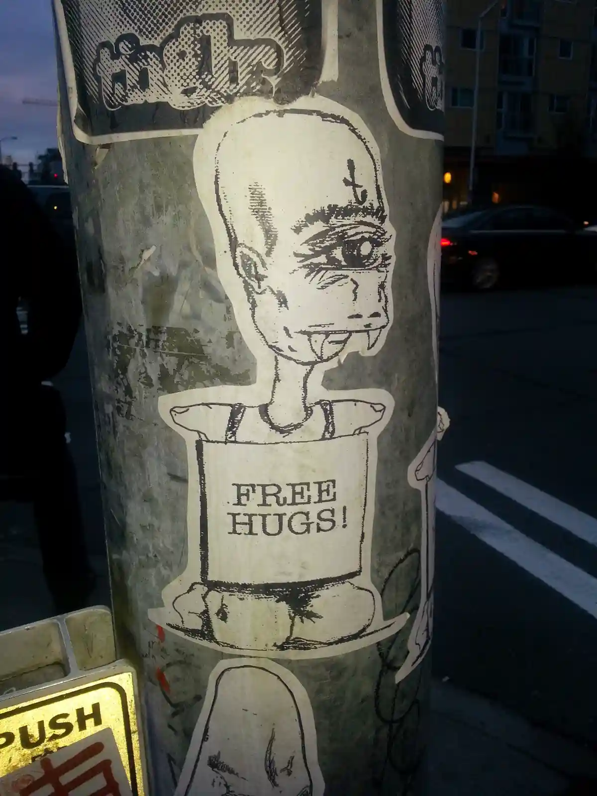 A black-and-white sticker on a street pole shows a cartoonish, bald figure with a cross on its forehead and fangs, wearing a…