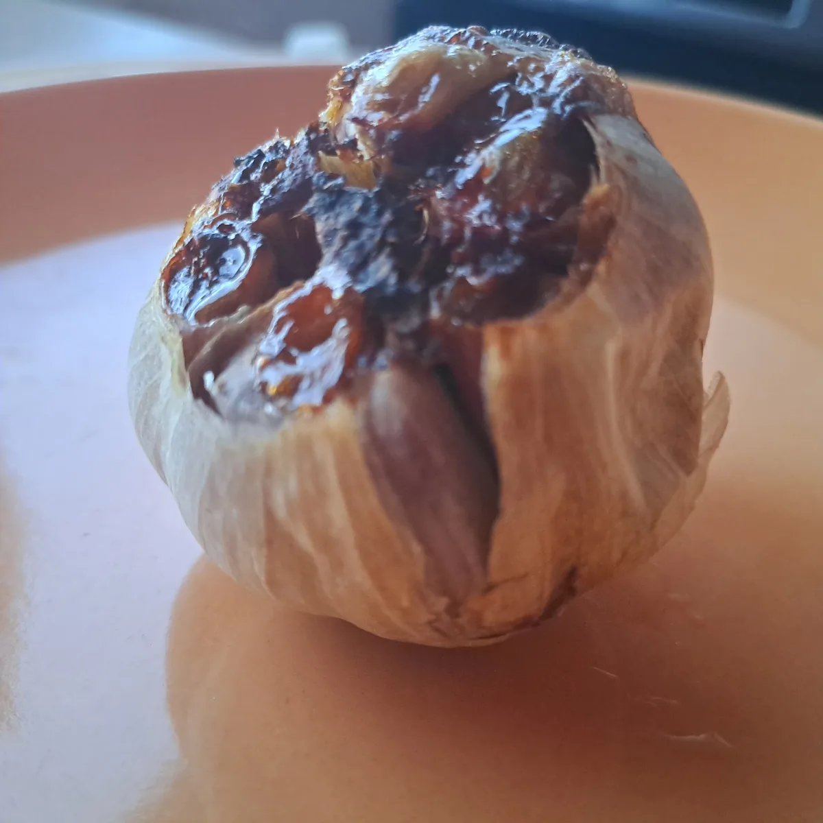 A close-up photo of a roasted garlic bulb sitting on a light orange plate, its top split open to reveal deeply caramelized…