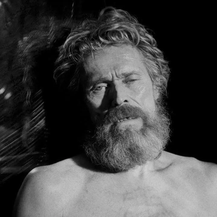A black-and-white photo shows a shirtless man with a full beard and wild hair, looking weary and slightly annoyed. The text…