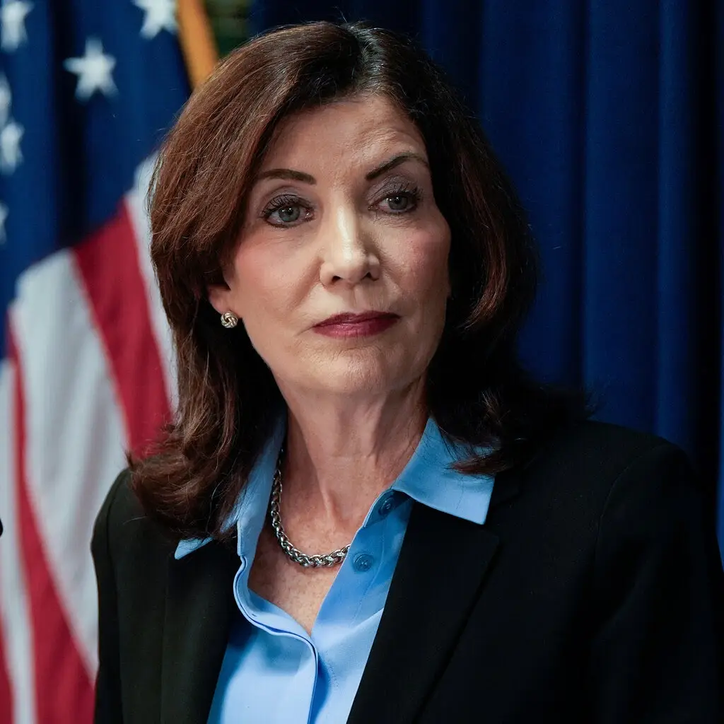 a woman with brown hair, wearing a blue shirt and black blazer, stands in front of an american flag and blue curtain. Her…