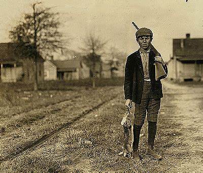 A sepia-toned historical photograph shows a young man in early 20th-century attire standing outdoors near train tracks…