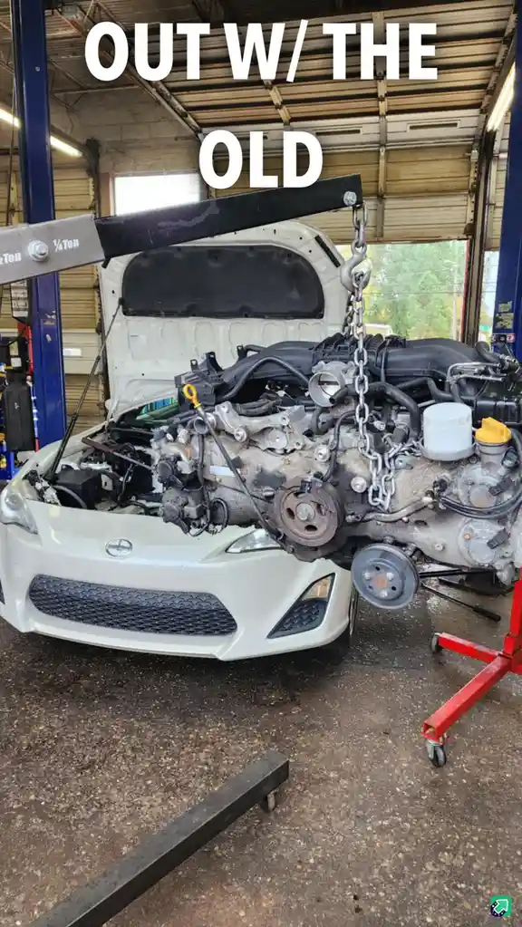 A white car with its hood open is in a garage, and its engine is being lifted out by a chain attached to a hoist. The…