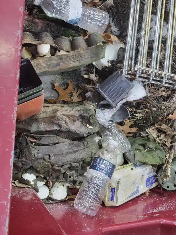 A red trash can is overflowing with discarded items including broken egg cartons with cracked eggs, plastic water bottles…