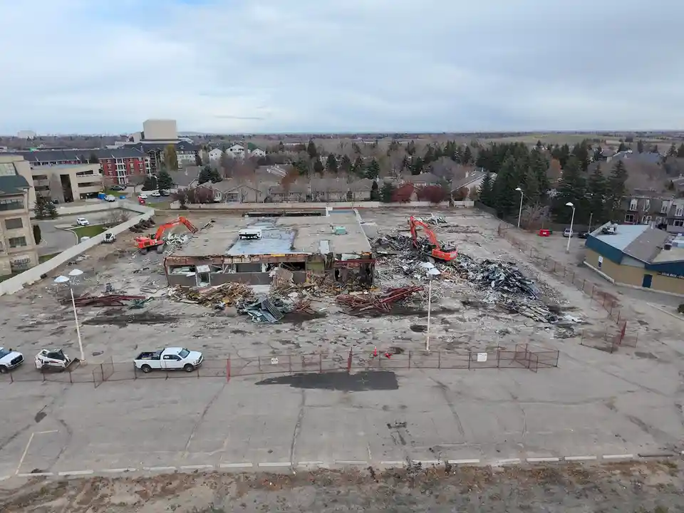 An aerial view shows a large building under demolition with two orange excavators tearing it down, leaving piles of rubble…