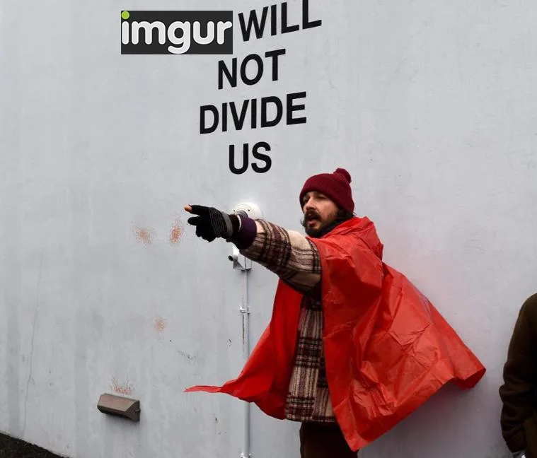 A man in a red poncho and beanie points emphatically at a white wall that has the text 'imgur WILL NOT DIVIDE US' painted on…