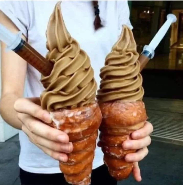 A person holds two large, spiral-shaped soft-serve ice cream cones made from a brown, coffee-colored frozen dessert, each…