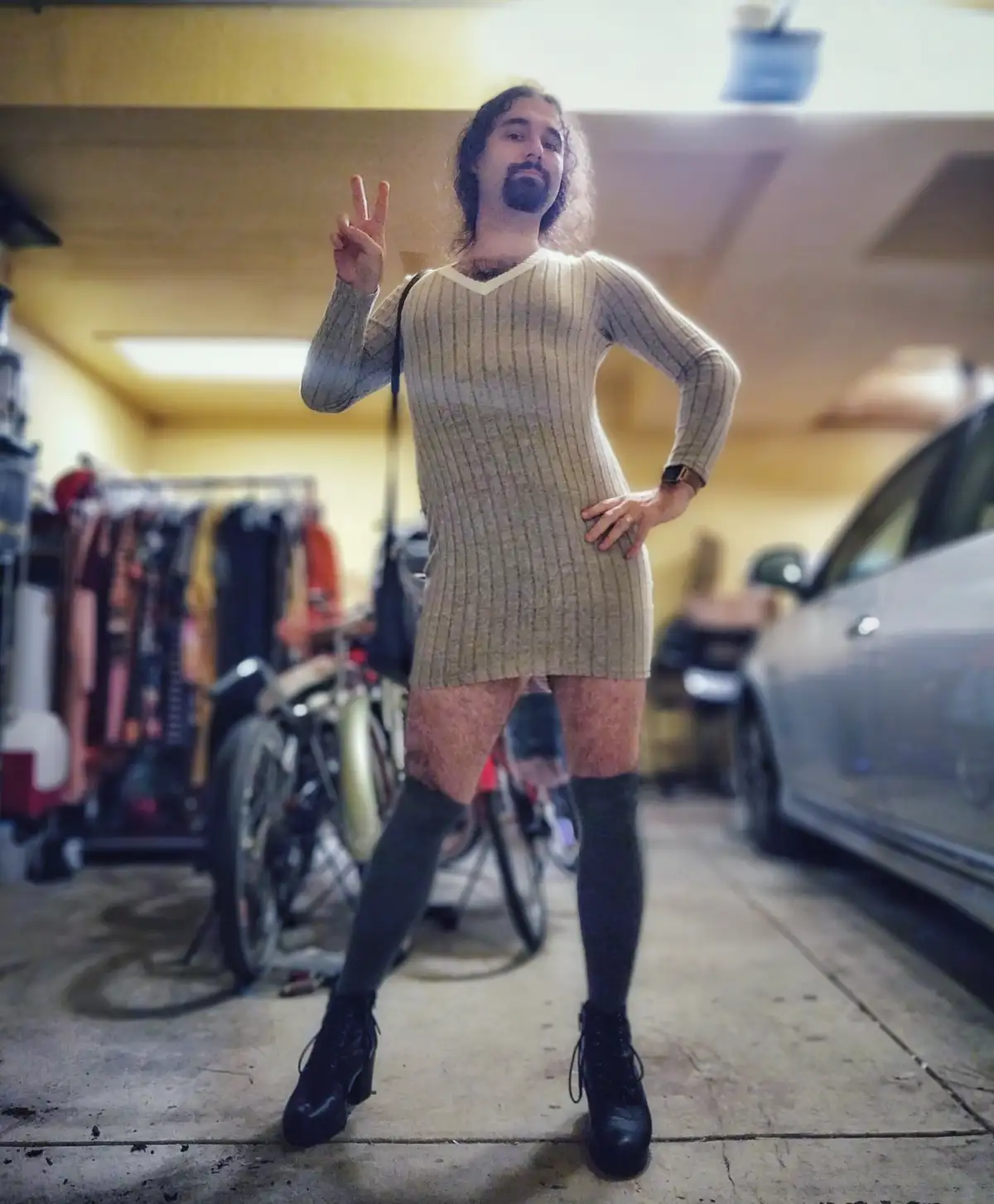 A man with long hair and a beard stands in a garage wearing a tight, ribbed, beige sweater dress, thigh-high black socks…