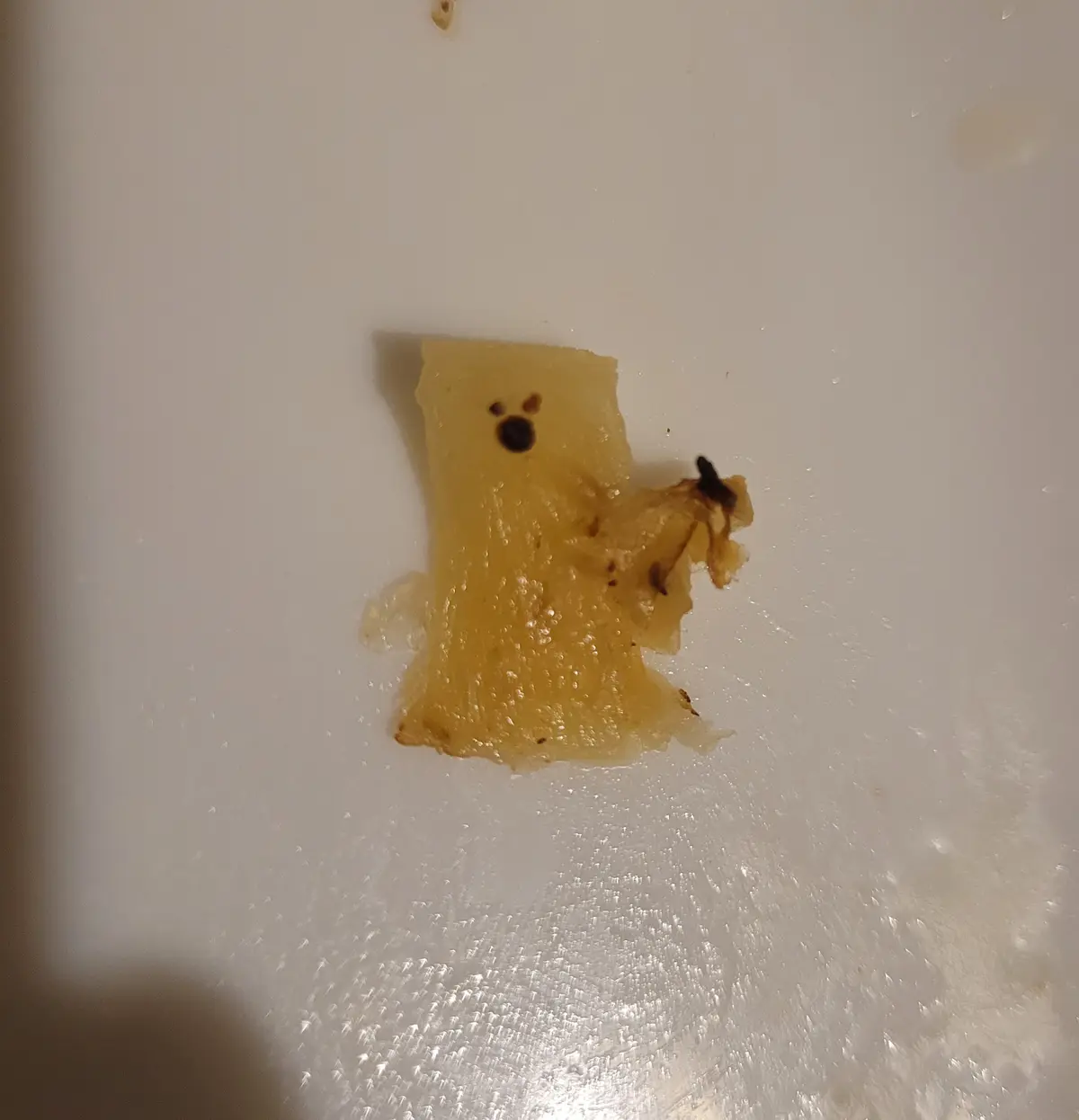 A small, translucent, yellowish piece of pineapple rests on a white textured surface, with dark spots resembling eyes and a…