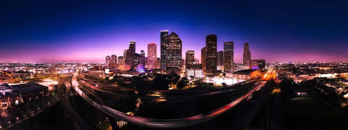 A panoramic night photograph of the Houston skyline with illuminated skyscrapers against a purple twilight sky, featuring…