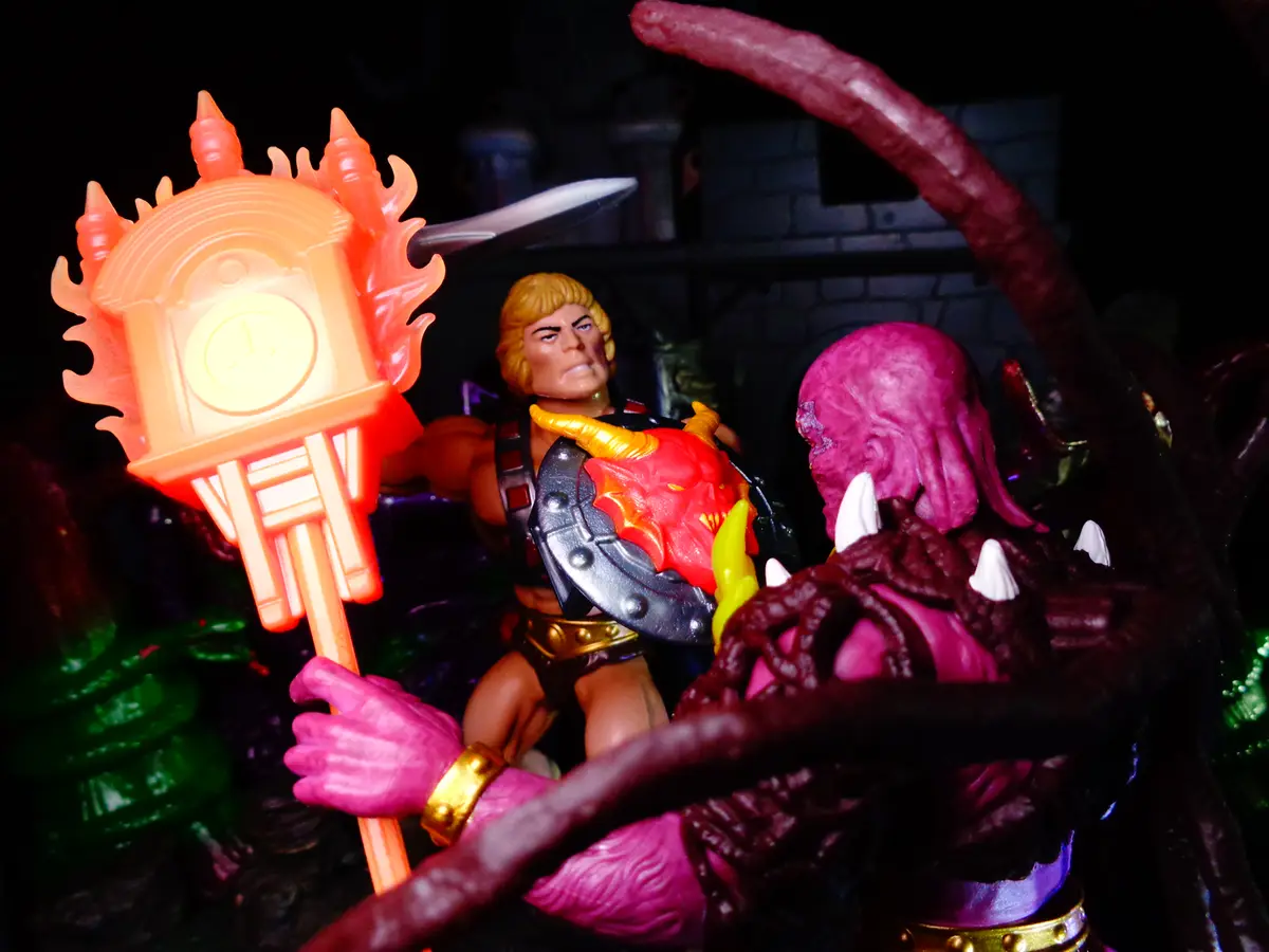 A toy He-Man figure with blonde hair and armor holds a glowing orange clock tower weapon while facing a pink-skinned…
