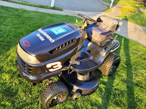 A black riding lawnmower is parked on a green lawn, modified to resemble a GM service vehicle with a red cross, 'Goodwrench…