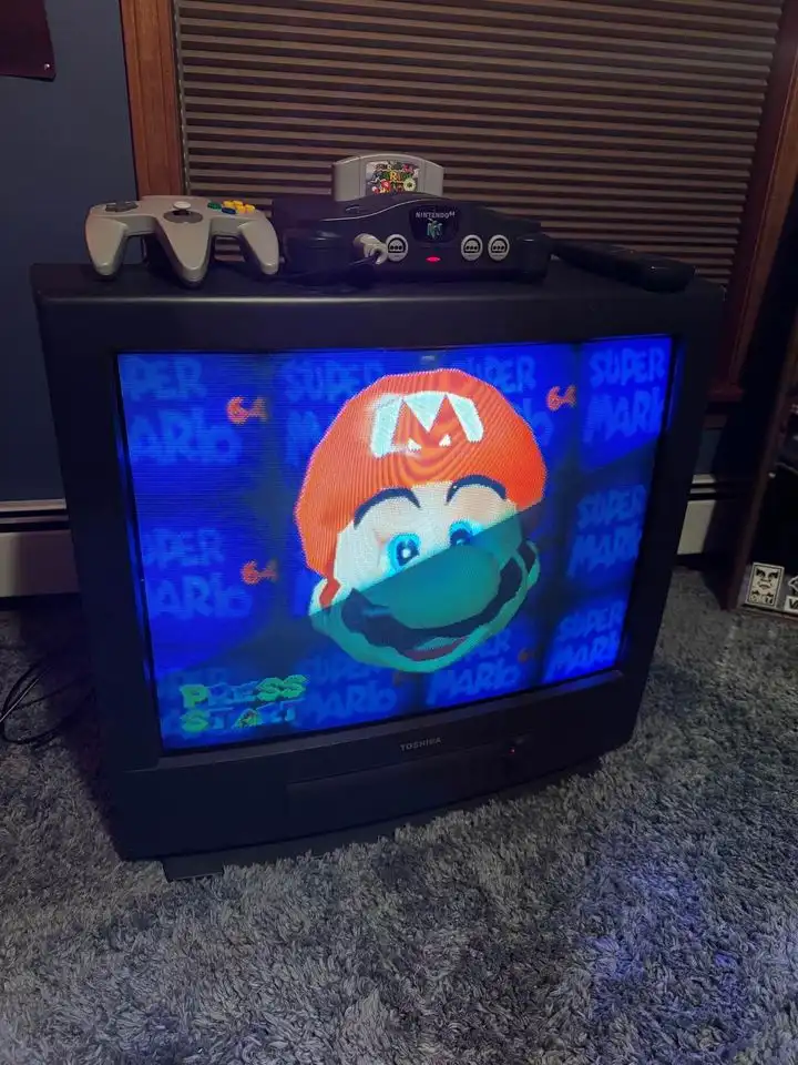 A CRT television displays the Super Mario 64 title screen with Mario's face, while a Nintendo 64 console and controller sit…