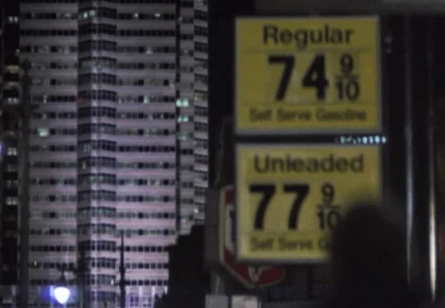 A blurry nighttime photo of a gas station price sign showing 'Regular 74 9/10' and 'Unleaded 77 9/10' with a tall lit-up…